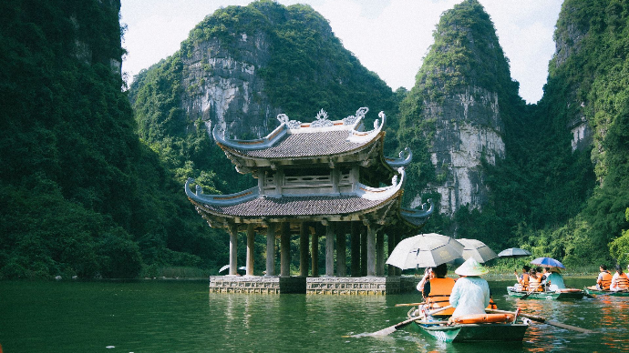 Temple sur l'eau au Vietnam avec en arrière plan des montagnes. Au premier plan, des personnes à bord de barques